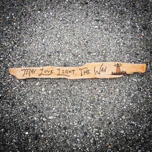 May include: A piece of driftwood with the words "May Love Light The Way" burned into it. A small lighthouse drawing is at the end of the wood.