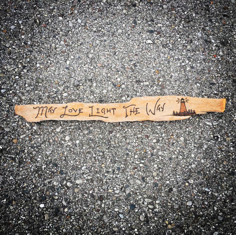 May include: A piece of driftwood with the words "May Love Light The Way" burned into it. A small lighthouse drawing is at the end of the wood.