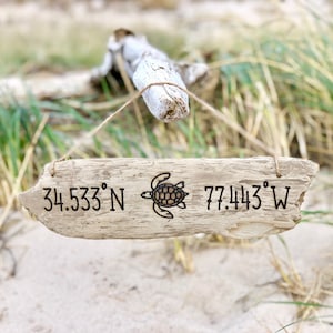 May include: A wooden sign with a sea turtle design and the coordinates 34.533° N 77.443° W. The sign is hanging from a piece of driftwood.