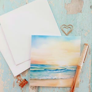 Puede incluir: Una pintura de acuarela de una escena de playa con olas azules y turquesas, una playa de arena y una puesta de sol. La pintura está junto a tarjetas blancas, un bolígrafo de oro rosa y clips. El fondo es una superficie de madera azul claro.