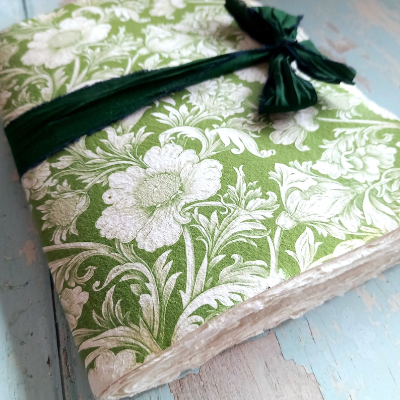 May include: A rectangular journal with a green and white floral pattern on the cover. The book is secured with a dark green ribbon tied in a bow. The pages have a rough, deckled edge texture, and the cover features a dense botanical print.