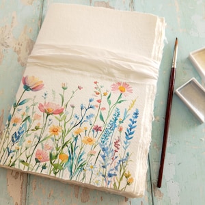 May include: A thick, off-white paper journal with deckled edges, wrapped in a white ribbon. The cover features a watercolour illustration of colourful wildflowers in shades of pink, blue, and yellow. A thin, dark brown paintbrush lies beside it.