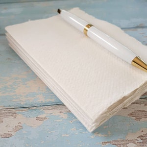 May include: A white notebook with a pen on top. The notebook is made of textured paper and has a rough edge. The pen is white with a gold trim.