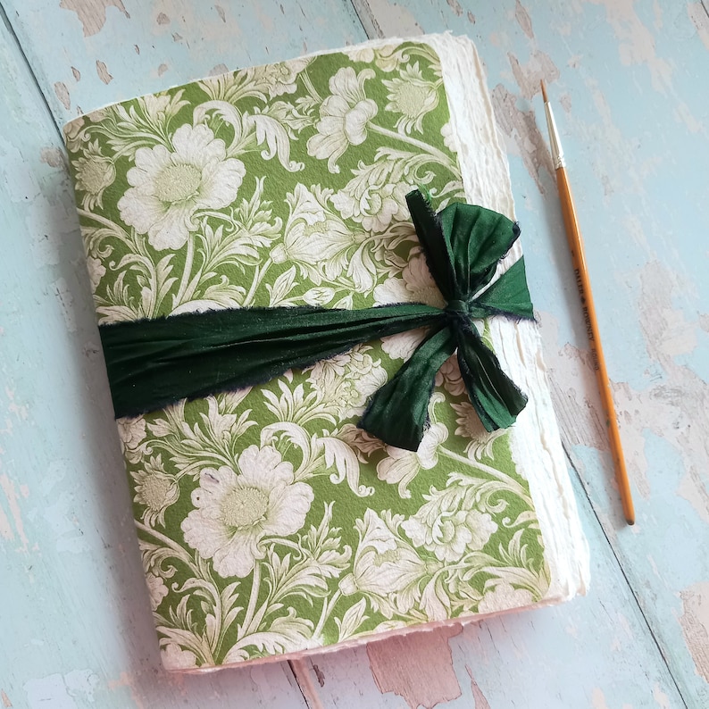May include: A journal with a green and white floral patterned cover, featuring a dark green fabric ribbon tied in a bow around the center. The journal has thick, textured, deckle-edged paper pages and is placed next to a thin wooden paintbrush.