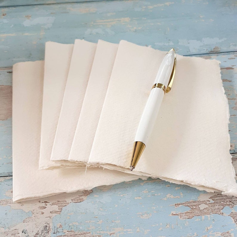 May include: A stack of four blank, white, handmade paper cards with deckled edges. A white pen with a gold trim is placed on top of the cards.