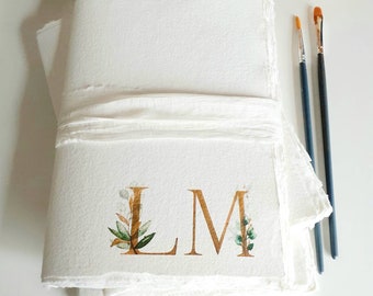 May include: A white, handmade journal with a gold ribbon and the initials "LM" in gold script with green watercolor floral accents.