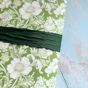 May include: A rectangular object covered in green paper with a white floral damask pattern. A wide, dark forest green ribbon with frayed, raw edges is wrapped horizontally across the center of the surface, creating a textured, layered appearance.