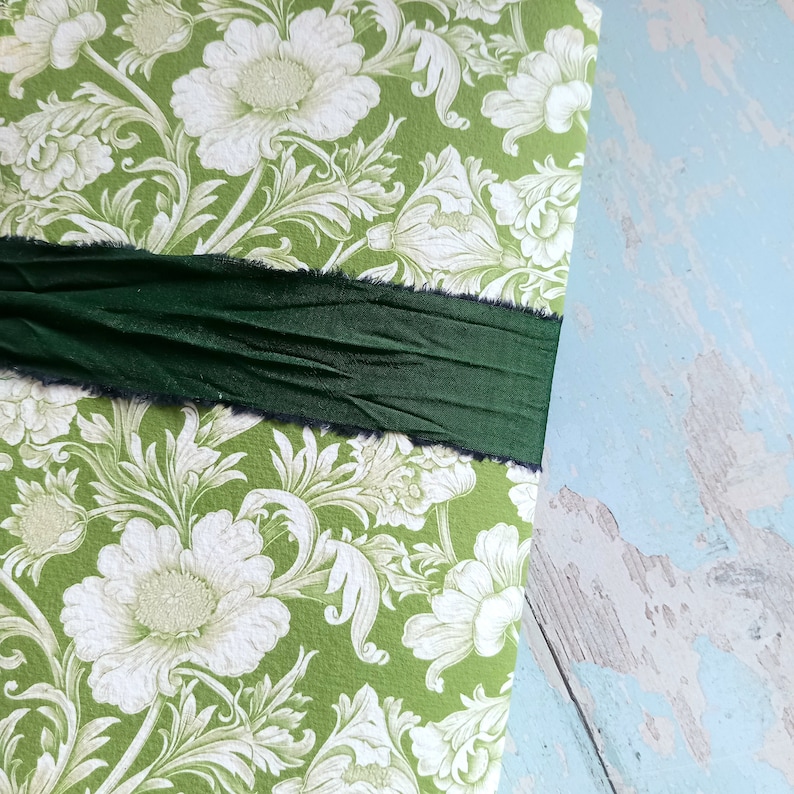 May include: A rectangular object covered in green paper with a white floral damask pattern. A wide, dark forest green ribbon with frayed, raw edges is wrapped horizontally across the center of the surface, creating a textured, layered appearance.