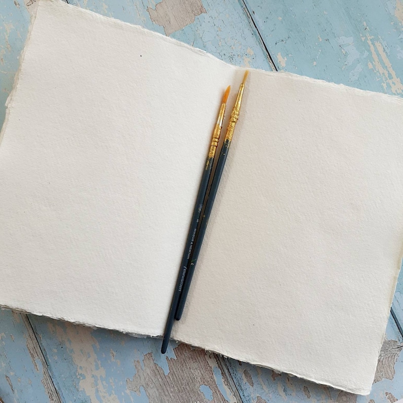 May include: An open sketchbook with thick, off-white, textured paper pages featuring deckled edges. Two dark-handled paintbrushes with gold-colored ferrules and light-colored bristles are placed diagonally across the center of the open pages.