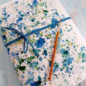 Può includere: Un quaderno bianco con schizzi di acquerello blu, verde e giallo. Un nastro blu è legato attorno al quaderno e un pennello di legno è appoggiato sul quaderno.