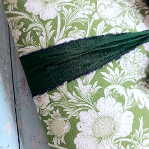 May include: A rectangular object covered in green paper featuring a white floral and leaf pattern. A dark forest green ribbon with frayed edges is wrapped horizontally across the center of the patterned surface, creating a textured contrast.
