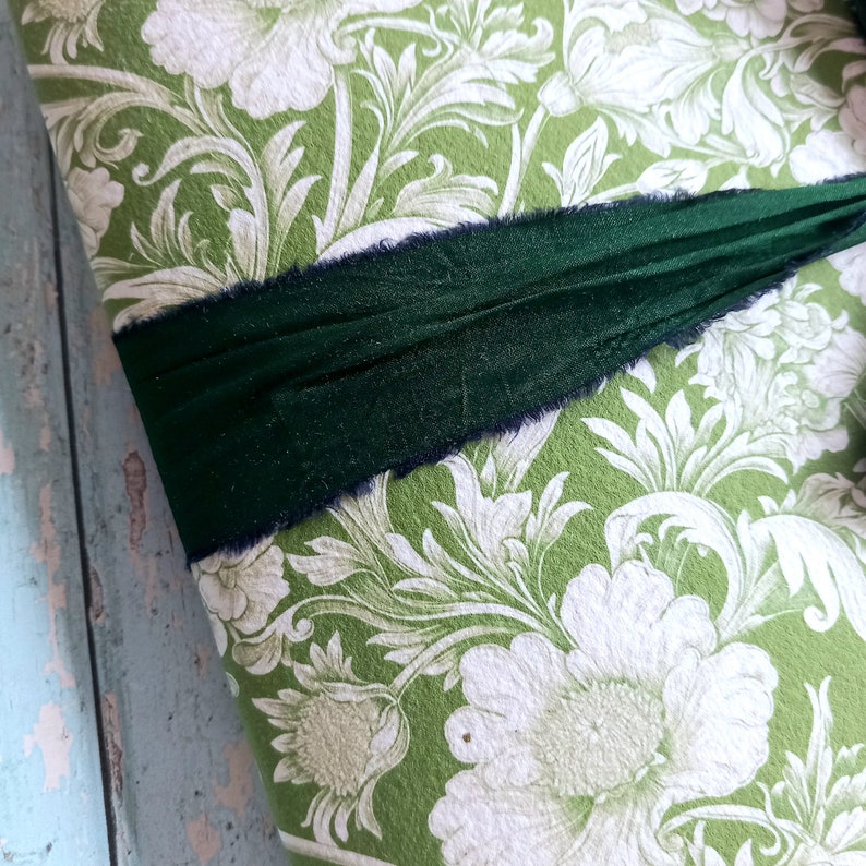 May include: A rectangular object covered in green paper featuring a white floral and leaf pattern. A dark forest green ribbon with frayed edges is wrapped horizontally across the center of the patterned surface, creating a textured contrast.