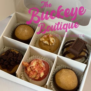 May include: A white box with six assorted gourmet sweets. The treats include chocolate-covered peanut butter balls, cookies with walnuts, and desserts topped with chocolate chips, Hershey's chocolate, and red sprinkles. The text "The Buckeye Boutique" is visible.