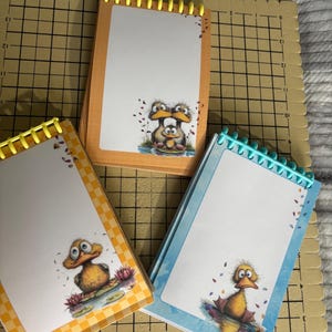 May include: Three spiral-bound notepads with duck illustrations. The notepads have orange, yellow, and blue borders, and each features a different duck character on the front. The notepads are on a beige macrame board.