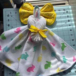 K&ouml;nnte beinhalten: Eine handgefertigte Osterhasen-Handtuchpuppe. Die Puppe besteht aus einem wei&szlig;en Handtuch mit bunten Hasenaufdrucken. Sie hat eine gelbe Schleife und eine gelbe Fliege. Der Kopf besteht aus einem gerollten wei&szlig;en Handtuch mit einem Regenbogenakzent.