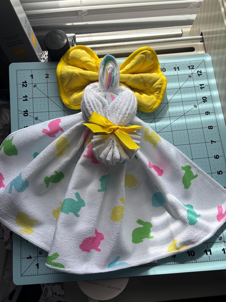 K&ouml;nnte beinhalten: Eine handgefertigte Osterhasen-Handtuchpuppe. Die Puppe besteht aus einem wei&szlig;en Handtuch mit bunten Hasenaufdrucken. Sie hat eine gelbe Schleife und eine gelbe Fliege. Der Kopf besteht aus einem gerollten wei&szlig;en Handtuch mit einem Regenbogenakzent.