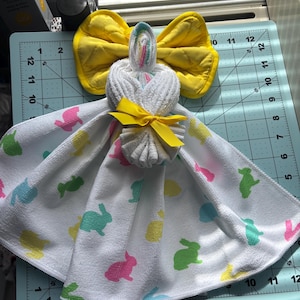 K&ouml;nnte beinhalten: Eine handgemachte Osterhasen-Handtuchpuppe. Die Puppe besteht aus einem wei&szlig;en Handtuch mit bunten Hasenaufdrucken. Sie hat eine gelbe Schleife f&uuml;r den Kopf und ein gelbes Band um den Hals. Die Puppe wird auf einer blauen Schneidematte ausgestellt.