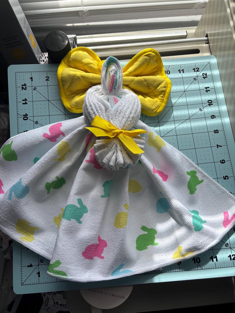 K&ouml;nnte beinhalten: Eine handgemachte Osterhasen-Handtuchpuppe. Die Puppe besteht aus einem wei&szlig;en Handtuch mit bunten Hasenaufdrucken. Sie hat eine gelbe Schleife f&uuml;r den Kopf und ein gelbes Band um den Hals. Die Puppe wird auf einer blauen Schneidematte ausgestellt.