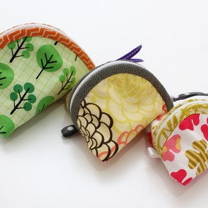 Pode incluir: Três bolsas de tecido com padrões diferentes. A primeira bolsa é verde com uma grade branca e uma borda laranja. A segunda bolsa é amarela com um padrão floral e uma borda cinza. A terceira bolsa é branca com um padrão floral rosa e amarelo e uma borda marrom.