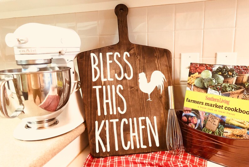 May include: A wooden cutting board with the words "BLESS THIS KITCHEN" and a rooster silhouette in white. A stainless steel whisk and a cookbook are also visible, suggesting a kitchen theme.