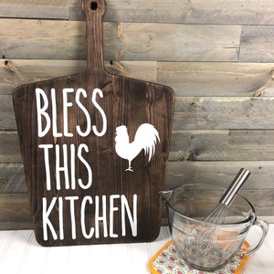 May include: A dark brown wooden cutting board with a handle, featuring the text "BLESS THIS KITCHEN" and a white rooster silhouette. A clear glass mixing bowl with a whisk and a patterned pot holder are next to the board.