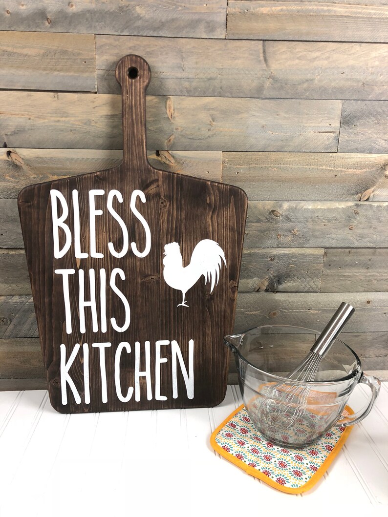 May include: A dark brown wooden cutting board with a handle, featuring the text "BLESS THIS KITCHEN" and a white rooster silhouette. A clear glass mixing bowl with a whisk and a patterned pot holder are next to the board.