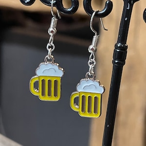 Peut inclure: Une paire de boucles d'oreilles pendantes argentées avec un charme de chope de bière jaune. La chope a un dessus de mousse blanc et est soulignée d'argent.
