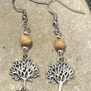 May include: A pair of silver-toned tree of life earrings. Each earring features a detailed tree charm, a light brown wooden bead, and a silver-colored ear wire. The earrings are designed for pierced ears and have a nature-inspired theme.