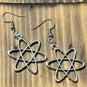 Könnte beinhalten: Ein Paar silberfarbene Ohrringe. Jeder Ohrring hat ein stilisiertes Atomdesign mit einem zentralen Kern und umlaufenden Elektronen. Die Ohrringe hängen an einem einfachen Haken und werden auf einer Holzoberfläche präsentiert.