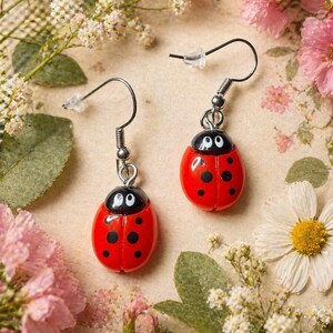 Pode incluir: Um par de brincos de joaninha. Cada brinco apresenta um corpo vermelho com manchas pretas e uma cabeça preta com olhos brancos. Os brincos têm ganchos prateados e são exibidos em um fundo floral.