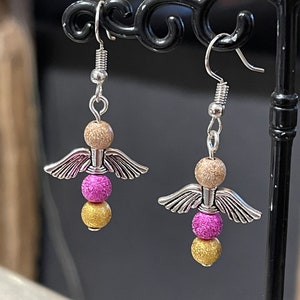 Pode incluir: Um par de brincos de asa de anjo prateados com três contas cada. As contas são douradas, cor-de-rosa e douradas.