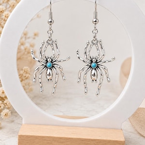 Op de afbeelding: Een paar zilverkleurige spinoorbellen met een metallic afwerking en een kleine, ronde, helderblauwe turkoois steen in het midden van het spinnenlichaam. De oorbellen hangen aan standaard zilverkleurige metalen oorhaakjes.