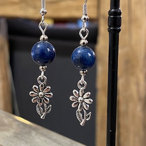 Peut inclure: Une paire de boucles d'oreilles pendantes en argent avec une perle ronde bleu foncé et un charme de fleur en argent.