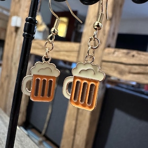 Peut inclure: Boucles d'oreilles dorées en forme de chope de bière. Chaque boucle d'oreille présente une chope orange avec de la mousse blanche. Elles sont suspendues à des crochets dorés, parfaites pour un look décontracté.
