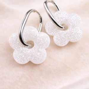 May include: A pair of white, glittery flower earrings. Each earring has a silver-coloured hoop attached to a flower-shaped charm. The earrings are presented on a soft, light-coloured background.