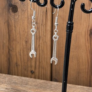 Puede incluir: Un par de pendientes plateados con forma de llave inglesa, colgados de un expositor negro. Los pendientes están diseñados con un diseño de llave inglesa, con un gancho y una cadena. El fondo es de madera.