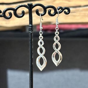 Peut inclure: Une paire de boucles d'oreilles pendantes argentées avec un design torsadé en forme d'infini. Les boucles d'oreilles sont suspendues à un support à boucles d'oreilles noir.