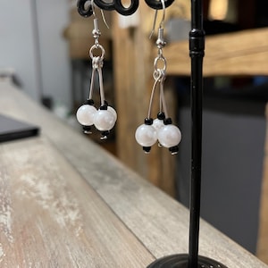 Può includere: Un paio di orecchini pendenti con tre perle bianche e perline nere su ogni orecchino. Gli orecchini sono appesi a un espositore per orecchini nero.