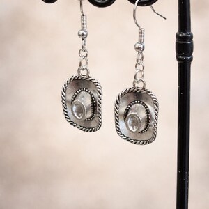 Peut inclure: Boucles d'oreilles argentées en forme de chapeau de cowboy. Chaque boucle d'oreille présente un petit pendentif en forme de chapeau détaillé avec une bordure en forme de corde et une pierre transparente de forme ovale au centre.