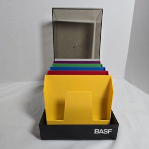 May include: A BASF branded black plastic desktop organizer featuring a series of tiered, upright slots in yellow, red, blue, green, and purple. The unit includes a tall, transparent, smoke-colored plastic lid that stands vertically at the back.