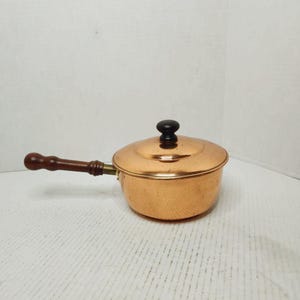 May include: A copper saucepan with a lid and a wooden handle. The handle is dark brown, and the lid has a black knob. The pan is round and has a shiny, reflective surface. The saucepan is designed for cooking.
