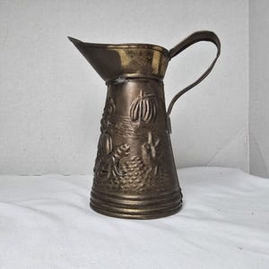 May include: A metallic bronze-toned pitcher featuring a tapered body and a curved handle. The exterior surface is decorated with embossed relief imagery depicting a sailing ship on water, a seated figure, and various textured landscape elements.