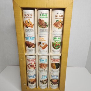 May include: A set of Bascom's seasoning tins in a cardboard display. The tins are white with various food images and labels like "Chicken," "Lamb," and "Steak." The display is a light brown color and holds the tins in neat rows.