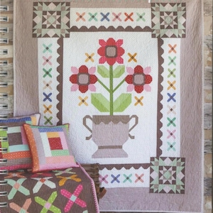 May include: A quilt with a floral design, pillows, and a small blanket. The quilt features a central image of flowers in a vase, surrounded by a border with cross and square patterns. The text "Prim & Proper" is at the top.