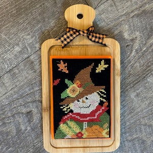 May include: A wooden cutting board with a black and orange checkered ribbon tied around the handle. A framed needlepoint design of a scarecrow with a brown hat and a red scarf is displayed on the board.