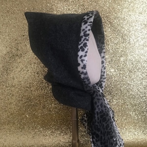 May include: A dark grey hooded scarf with a leopard print trim and scarf detail. The scarf is displayed on a mannequin head and stand against a gold glitter background. The hood is made of a soft, textured material.