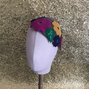 May include: A grey felt headband decorated with vibrant felt flowers in purple, yellow, and blue. Green felt leaves with gold detailing are also present. The headband is displayed on a mannequin head.