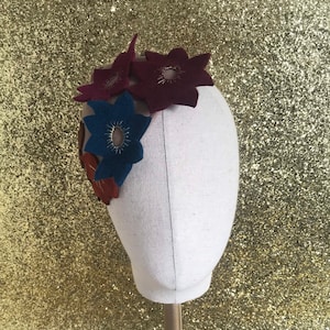 May include: A felt flower headband with three flowers in shades of burgundy, blue, and orange. The flowers have gold accents.