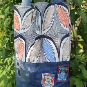 May include: A blue denim tote bag with a zipper closure. The bag has a patterned fabric lining with geometric shapes in shades of grey, blue, and orange. The bag has two denim straps and features three patches with the names "Cumberland", "Snowdonia", and "Snowdonia National Park" on the front.