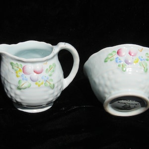May include: A light blue creamer and sugar bowl set. The creamer has a handle and spout, while the bowl is open. Both pieces have a textured surface and are decorated with a floral pattern in pink, yellow, and blue.